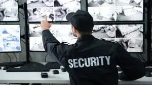 Guardia de seguridad sentado frente a múltiples monitores encendidos señalando uno, revisando la seguridad perimetral del área.