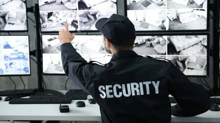 Guardia de seguridad sentado frente a múltiples monitores encendidos señalando uno, revisando la seguridad perimetral del área.