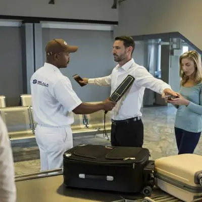 Guardia de seguridad revisa a una persona con un detector de metal, frente a una cinta transportadora de equipaje en un aeropuerto.